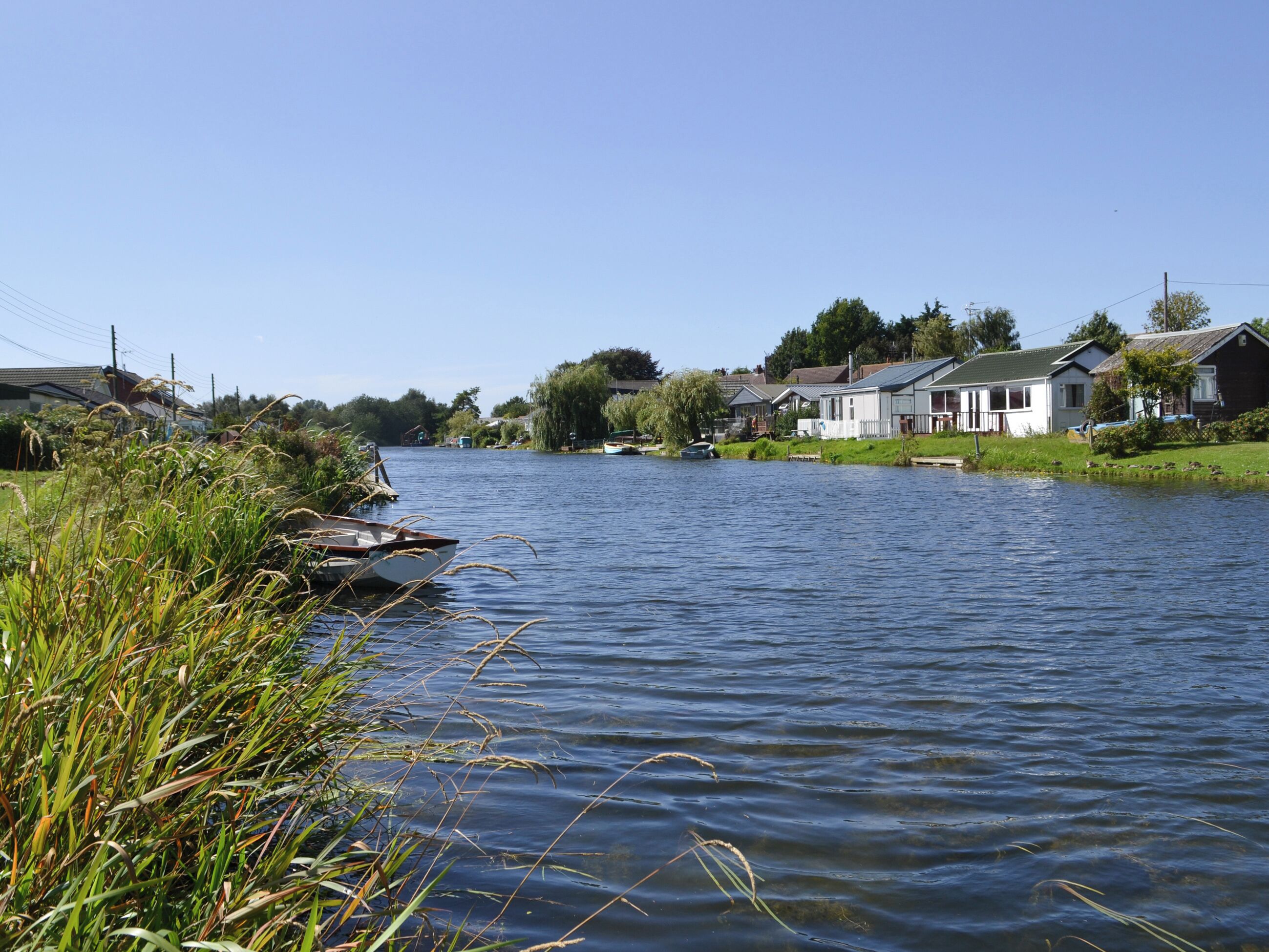 Un río tranquilo con barcas amarradas a la izquierda, rodeado de casas bajo un cielo despejado.