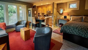Habitación de hotel moderna con una cama grande, un escritorio, dos sillas, una pequeña cocina y vistas al exterior a través de amplios ventanales.