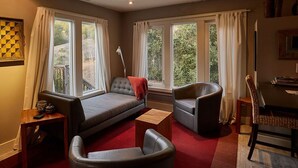 Una sala de estar acogedora con un sofá, dos sillones, una mesita de madera y una zona de comedor con una silla. Amplias ventanas ofrecen luz natural y vistas al exterior.