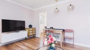 A modern living room with a flat-screen TV, a wooden dining table, a pink chair, and a glass coffee table with flowers.
