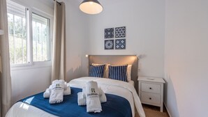 Une chambre d’hôtel comprenant un lit, deux oreillers, une table de chevet, une commode et un mur orné de quatre carreaux encadrés.