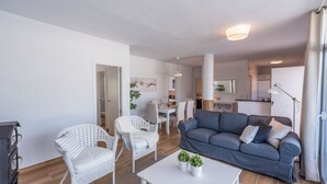 Un salon moderne avec un canapé gris, des fauteuils en osier blancs, une table basse blanche et, au fond, un coin repas.