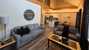 A spacious living area with a high ceiling, a wooden staircase, a kitchen in the background, and a large wall clock.