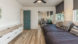 A modern bedroom with a wooden floor, a bed with multiple pillows, a ceiling fan, and a window with a view.