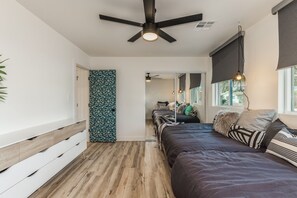 A modern bedroom with a wooden floor, a bed with multiple pillows, a ceiling fan, and a window with a view.
