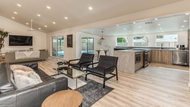 A modern living room with a sofa, chairs, a coffee table, and a TV. There is a kitchen area with a stove, oven, and refrigerator.