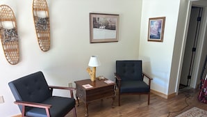 Una habitación con dos sillones oscuros, una mesita de madera con una lámpara y dos cuadros en la pared.