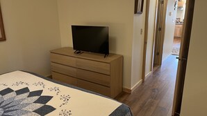 Un dormitorio con cama, un aparador con televisor, un pasillo que conduce a una cocina y una puerta a otra habitación.