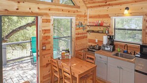 A wooden cabin with a dining table and chairs, a kitchen with a sink and stove, and large windows offering views of the outdoors. - Luxury Treehouse w/ Grill, Fire Pit, & Pond in Dripping Springs (Dripping Springs)