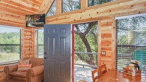 A cozy cabin interior with a wooden ceiling, a gray door, a cushioned chair, a wooden table, and a window overlooking greenery. - Luxury Treehouse w/ Grill, Fire Pit, & Pond in Dripping Springs (Dripping Springs)