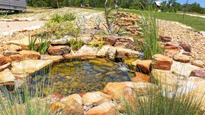Un petit étang entouré de rochers et de plantes, avec une terrasse en bois et un kiosque en arrière-plan.