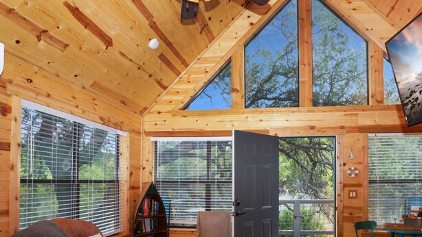 Interior de una cabaña de madera con una puerta grande abierta que da a una terraza exterior, una estantería, un sillón y una mesa con vistas a árboles a través de las ventanas.