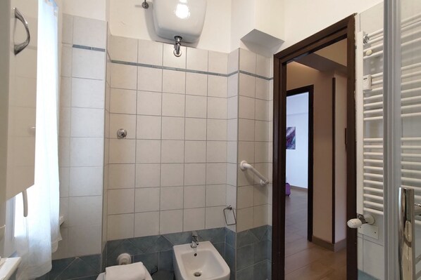 Un baño con inodoro, bidé y lavabo, paredes revestidas de azulejos y una puerta que da a otra habitación.