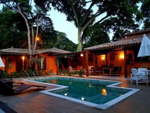 Un espace piscine avec des chaises longues et un coin repas, le tout entouré d’arbres et d’un bâtiment doté d’un toit en tuiles.