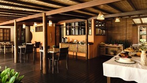 Une salle à manger spacieuse, dotée d’un plancher en bois et d’un plafond haut. On y trouve plusieurs tables à manger avec des chaises, et on aperçoit une cuisine à l’arrière-plan.