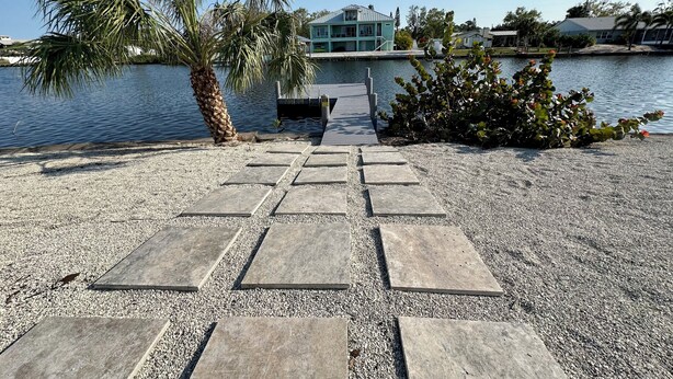 Un sendero de piedras guía hacia un muelle a orillas de un lago tranquilo, con una palmera y un arbusto de flores rojas cerca.