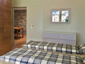 A bedroom with a bed, a dresser, a door leading to another room, and a window with a view of buildings.