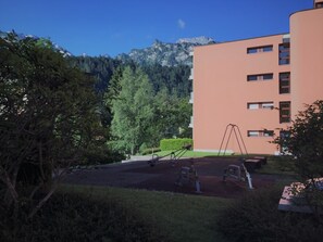 A playground with swings and a seesaw, surrounded by trees and a building with multiple windows.