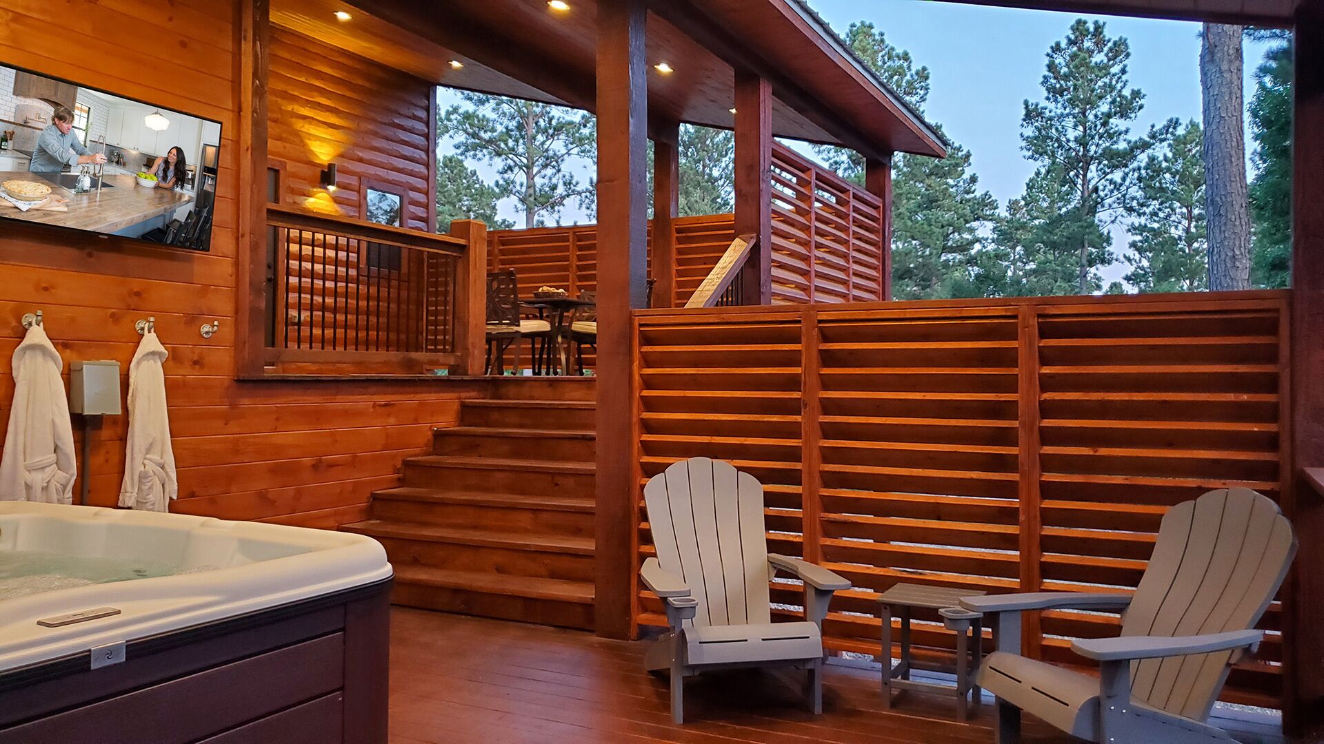 A cabin with a hot tub, wooden deck, Adirondack chairs, and a staircase leading to an upper level.