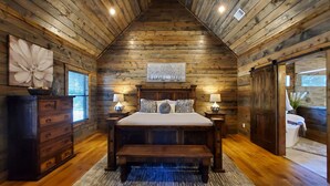 Un dormitorio de cabaña de troncos con techo de madera, una cama con dos almohadas, una mesita de noche con una lámpara y un aparador.