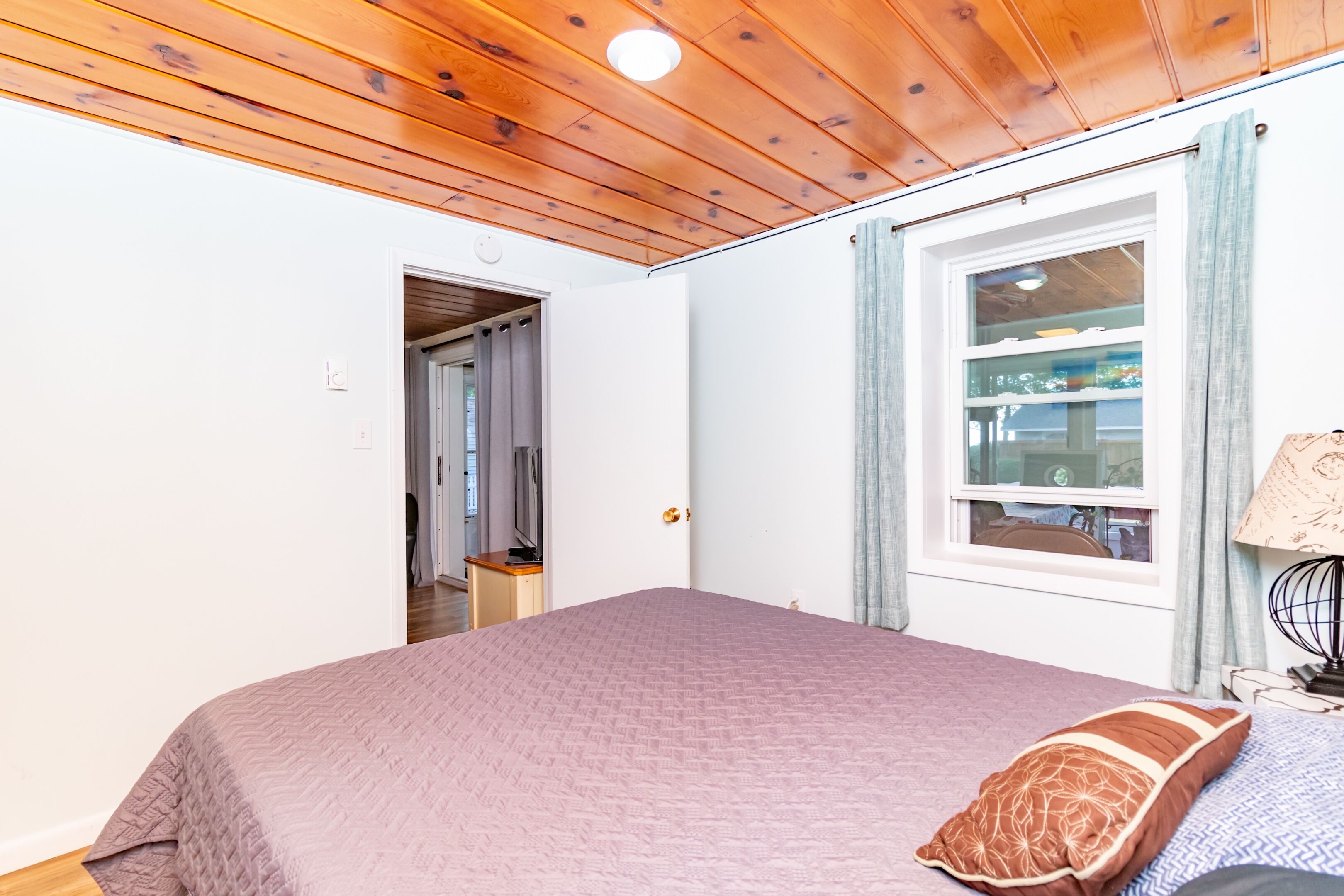 Ein Schlafzimmer mit Holzbalkendecke, einem Bett mit rosa Quilt, einem Fenster mit Vorhängen und einer Lampe auf einem Nachttisch.