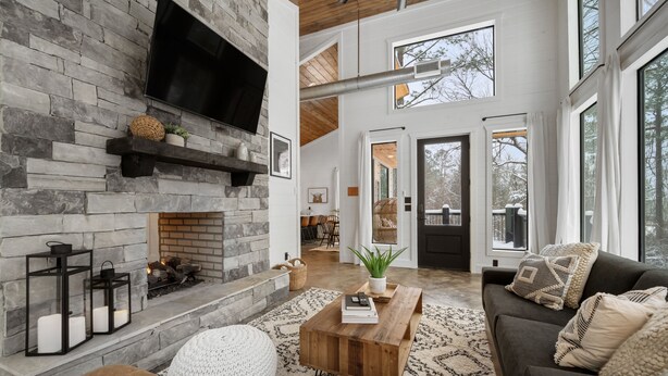 A modern living room with a stone fireplace, a large window, and a cozy seating area. - Luxury Honeymoon/Small Family, Hot Tub, Firewood Included (Broken Bow)