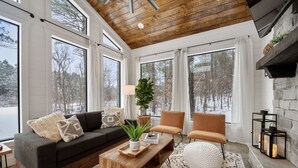 A cozy living room with a wooden ceiling, large windows, a fireplace, and a stone wall. - Luxury Honeymoon/Small Family, Hot Tub, Firewood Included (Broken Bow)