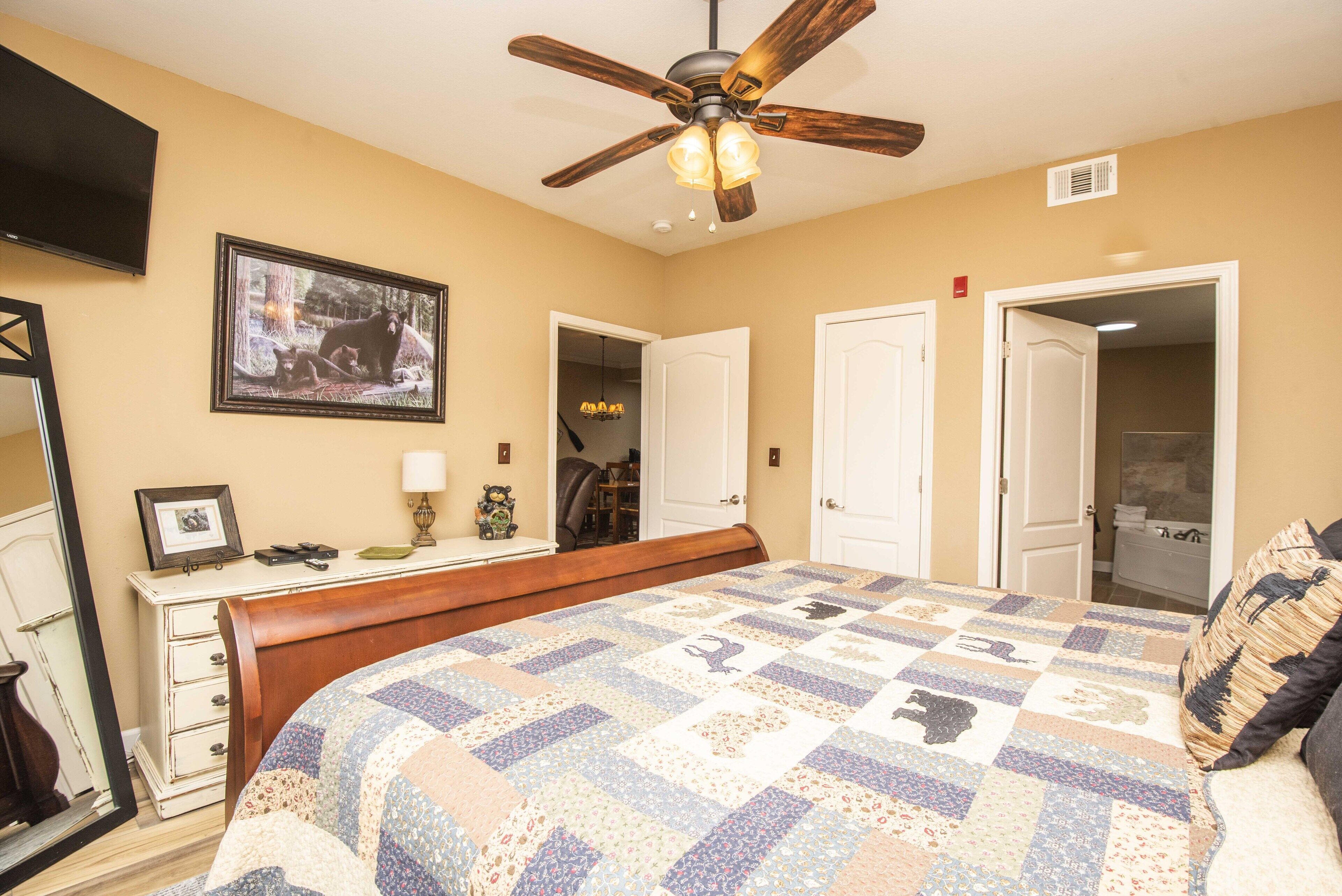 A bedroom with a bed, a ceiling fan, a dresser, a mirror, a framed picture on the wall, and a door leading to another room.