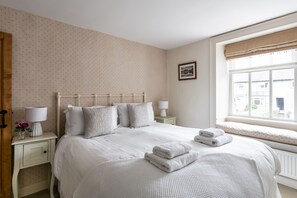 A neatly made bed with white linens and pillows, a bedside table with a lamp and a vase of flowers, and a window with a view of buildings outside. - Maggie Puddle Cottages (Grange-over-Sands)