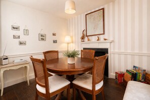 A dining room with a round wooden table, four wooden chairs, a lamp, a fireplace, and a bookshelf with books. - Maggie Puddle Cottages (Grange-over-Sands)