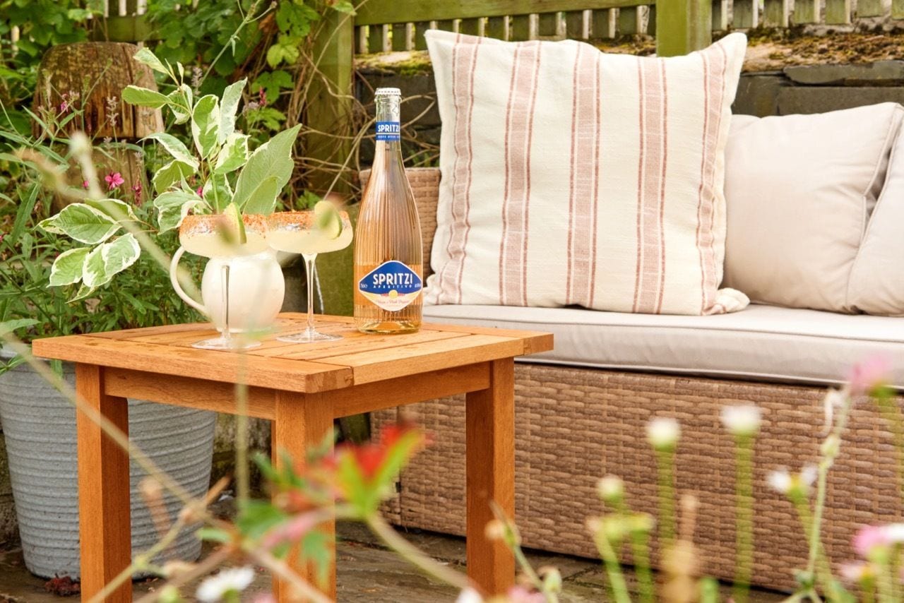 A wooden table with a bottle of Spritz, a glass, and a pitcher of a beverage, set on a patio with a wicker sofa and striped pillows.