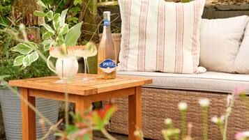 A wooden table with a bottle of Spritz, a glass, and a pitcher of a beverage, set on a patio with a wicker sofa and striped pillows.