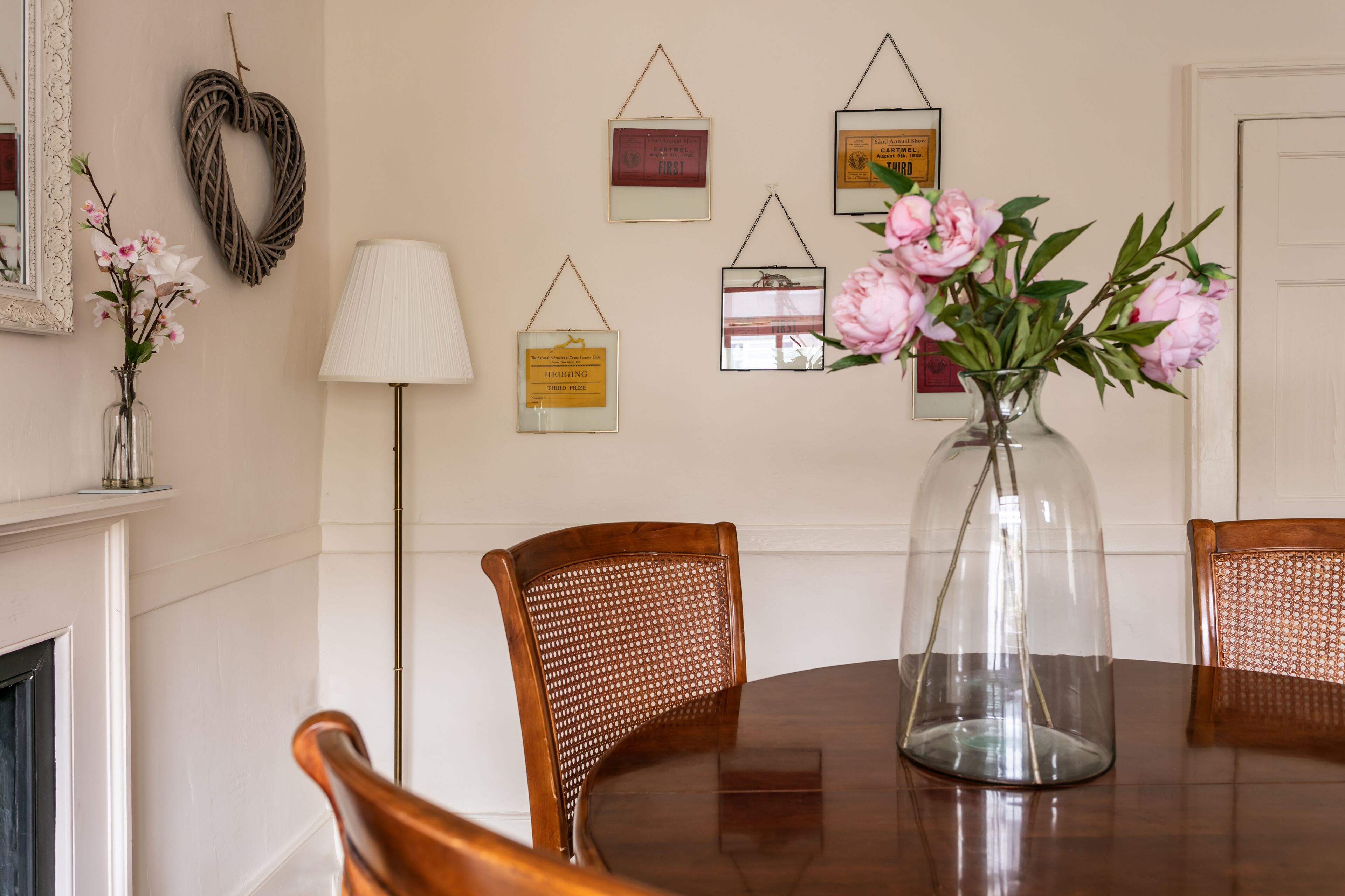 Une salle à manger avec une table ronde en bois, deux chaises en osier, un vase avec des fleurs et des décorations murales, notamment une couronne en forme de cœur et des certificats encadrés.