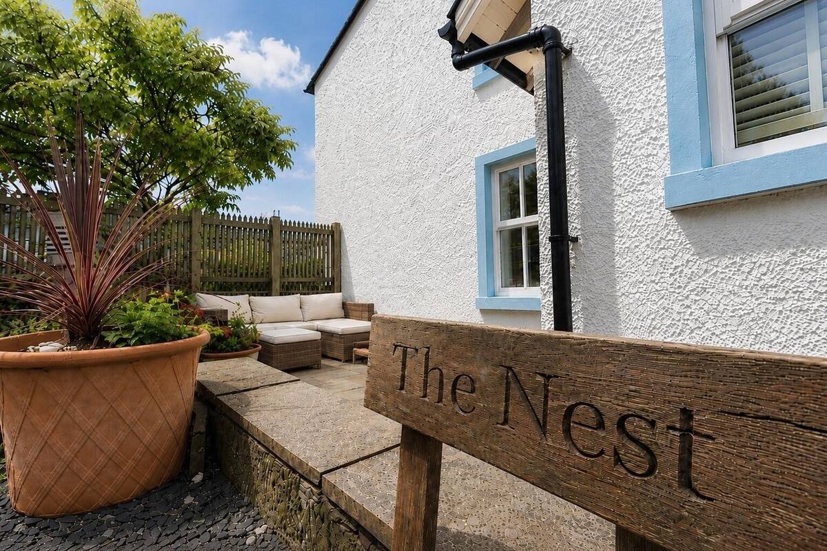 A wooden bench with 'The Nest' engraved on it, a white building with blue trim, and a wooden fence in the background.