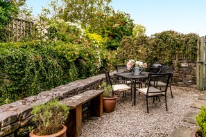 A patio with a stone bench, a table, and chairs, surrounded by lush greenery and a wooden fence. - Maggie Puddle Cottages (Grange-over-Sands)