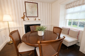 A dining room with a round wooden table, four wooden chairs, a potted plant, a fireplace, and a window with a view of greenery. - Maggie Puddle Cottages (Grange-over-Sands)