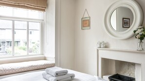 A neatly arranged bedroom with a large window, a fireplace, and a round mirror on the wall.