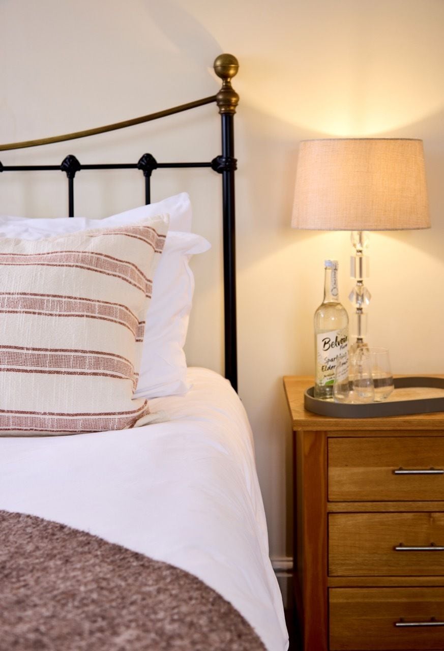 Ein Bett mit schwarzem Metallgestell, weißer Bettwäsche und gestreifteren Kissen. Ein Nachttisch mit einer Lampe, einer Flasche und Gläsern ist sichtbar.