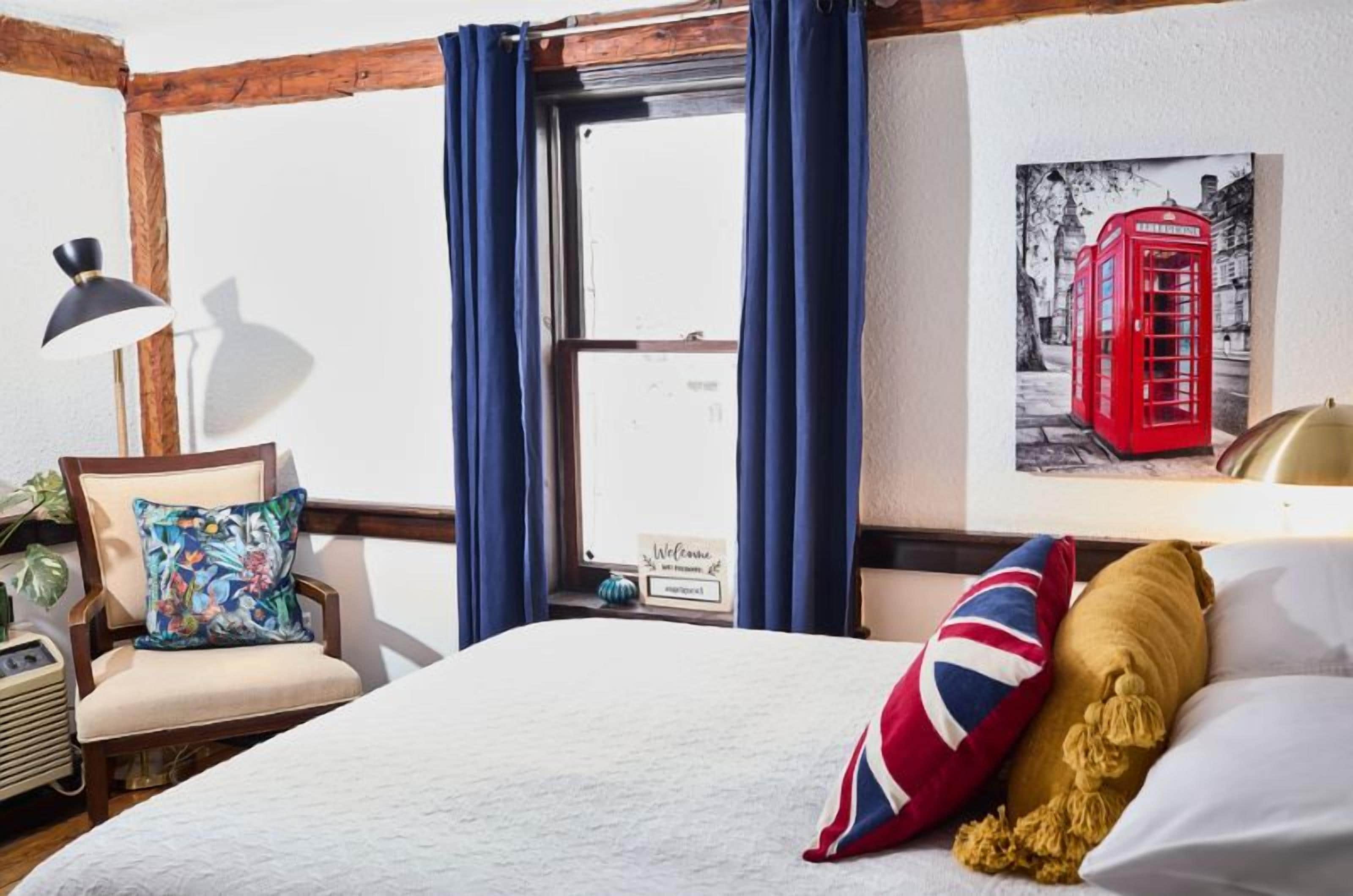 Una habitación de hotel con una cama grande, una silla con un cojín de colores, una lámpara y un cuadro con una cabina telefónica roja en la pared.
