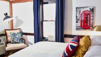 Una habitación de hotel con una cama grande, una silla con un cojín de colores, una lámpara y un cuadro con una cabina telefónica roja en la pared.