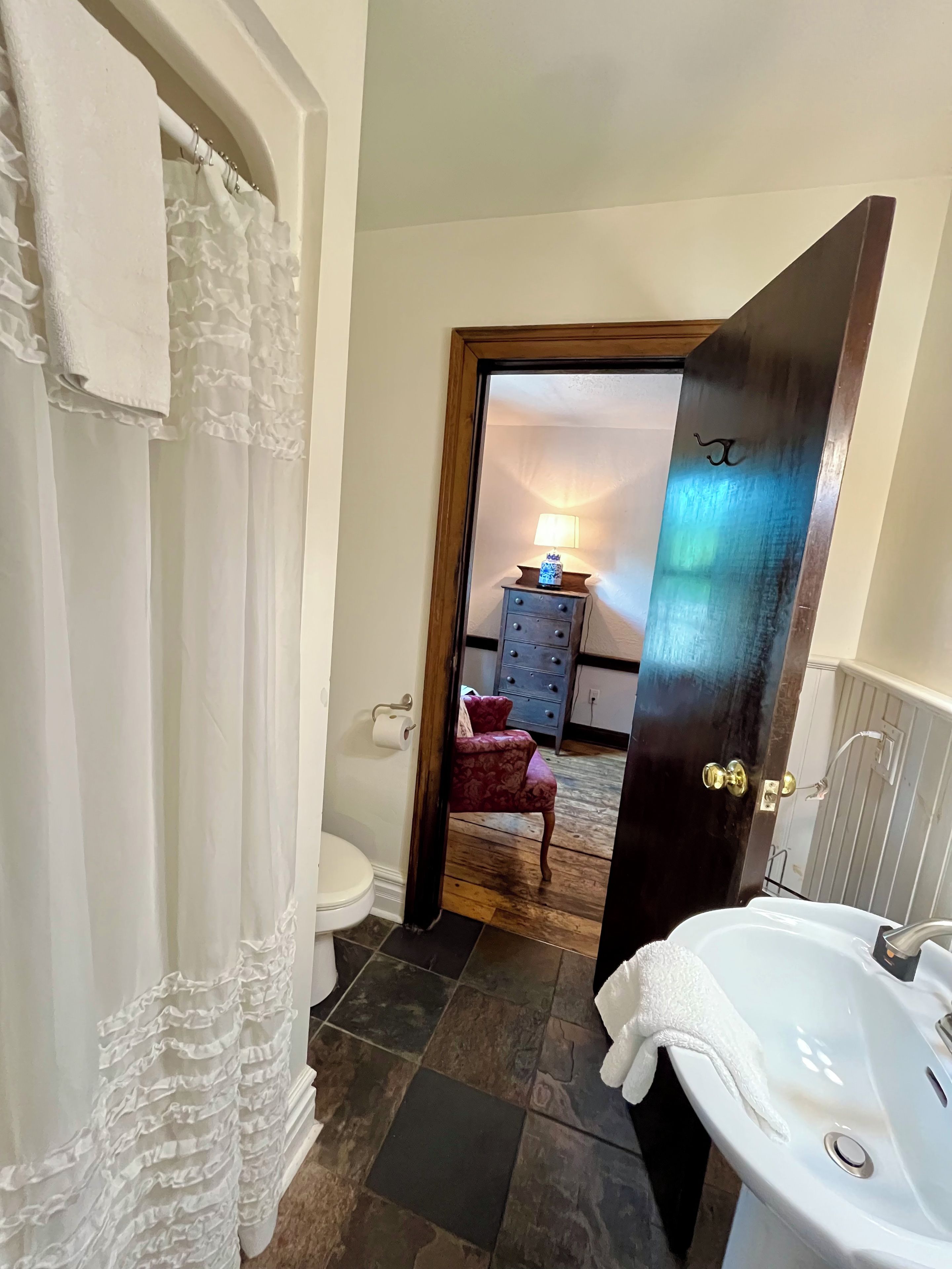 Une salle de bain avec une baignoire blanche, des toilettes, un miroir et une porte donnant sur une chambre à coucher équipée d’une commode et d’une lampe.
