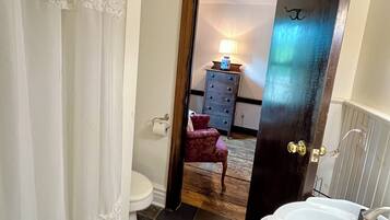 Une salle de bain avec une baignoire blanche, des toilettes, un miroir et une porte donnant sur une chambre à coucher équipée d’une commode et d’une lampe.