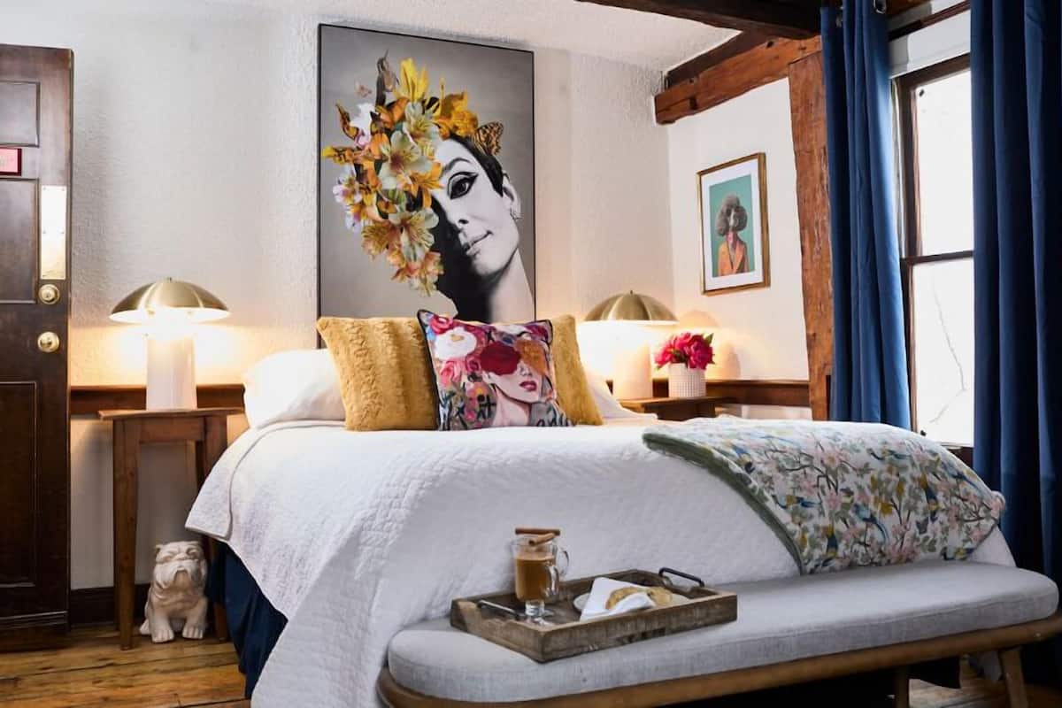 A well-appointed bedroom with a large bed, floral pillows, a bedside table with a lamp, a small statue, and a framed artwork on the wall.