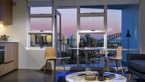 Una sala de estar moderna con vista a la ciudad, una mesa redonda con libros y un jarrón, y un ventanal con vistas a rascacielos y un puente.