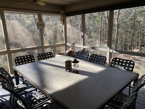 A covered outdoor dining area with a table and chairs, large windows, and a view of trees.