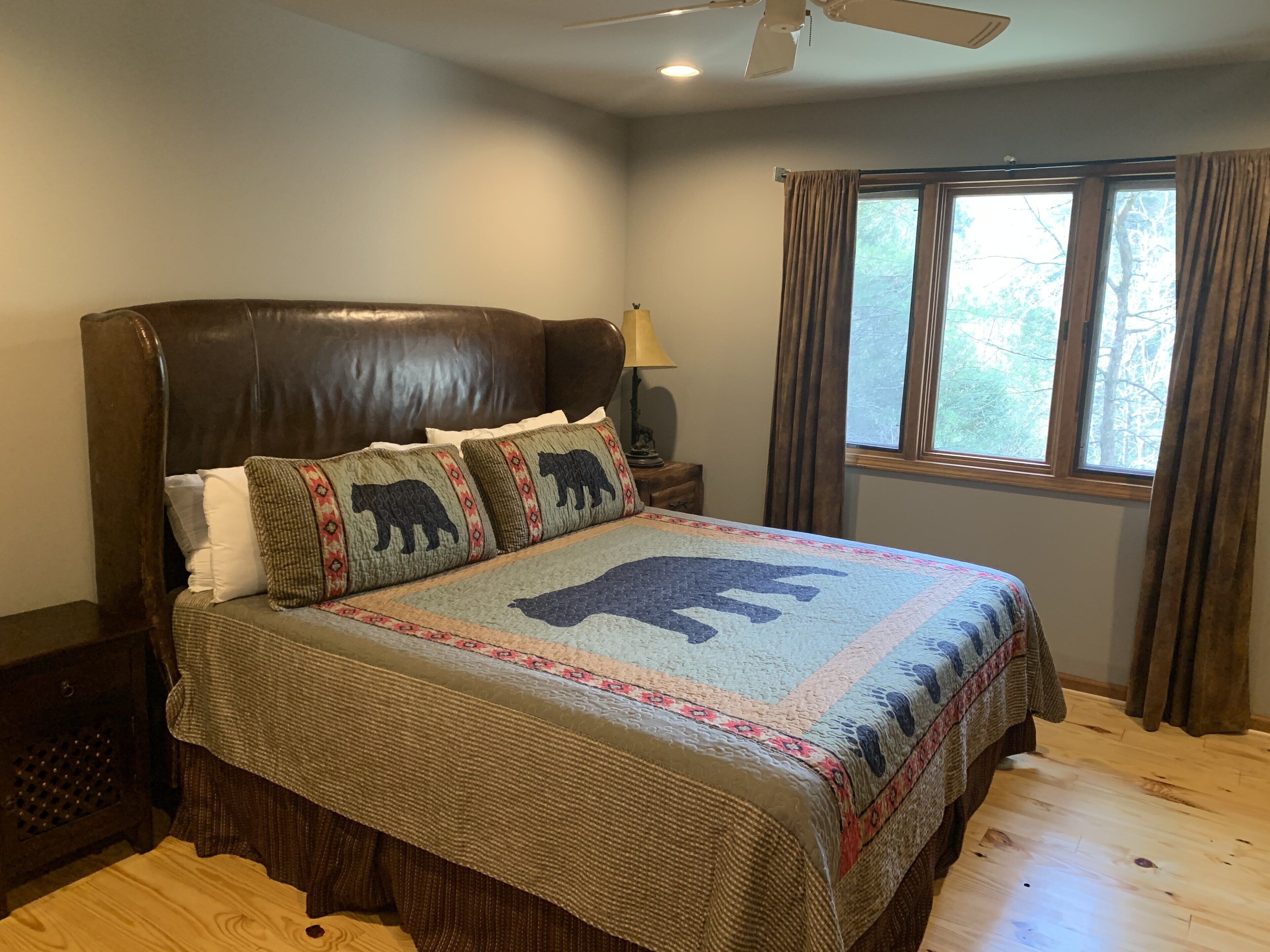 A bedroom with a large bed, a ceiling fan, a window with curtains, and a nightstand with a lamp.