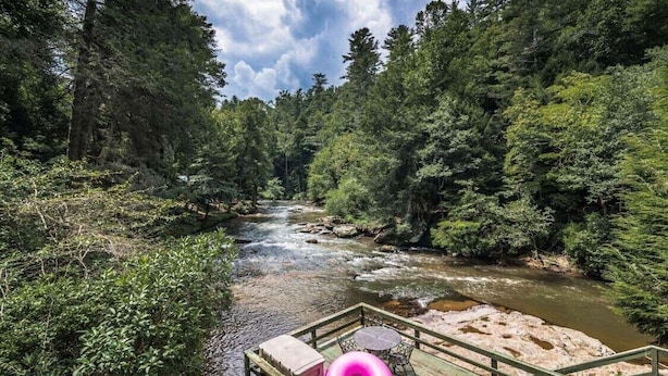 A river flowing through a forested area with a wooden deck and seating area in the foreground.