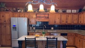 A kitchen with wooden cabinets, a granite countertop island, two bar stools, a refrigerator, and a chandelier.