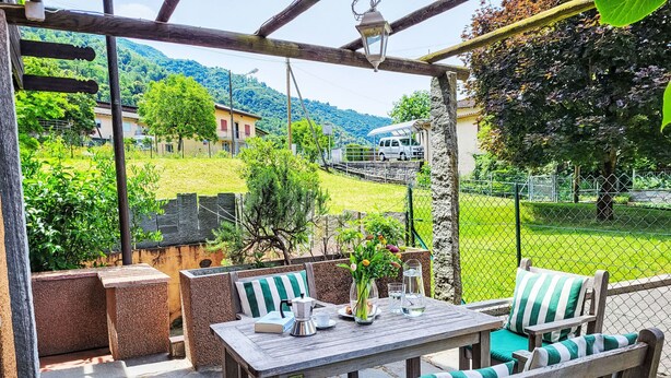 An outdoor patio with a wooden table and chairs, a view of a green landscape, and a stone wall. - Fiorita by Interhome (Brusino Arsizio)
