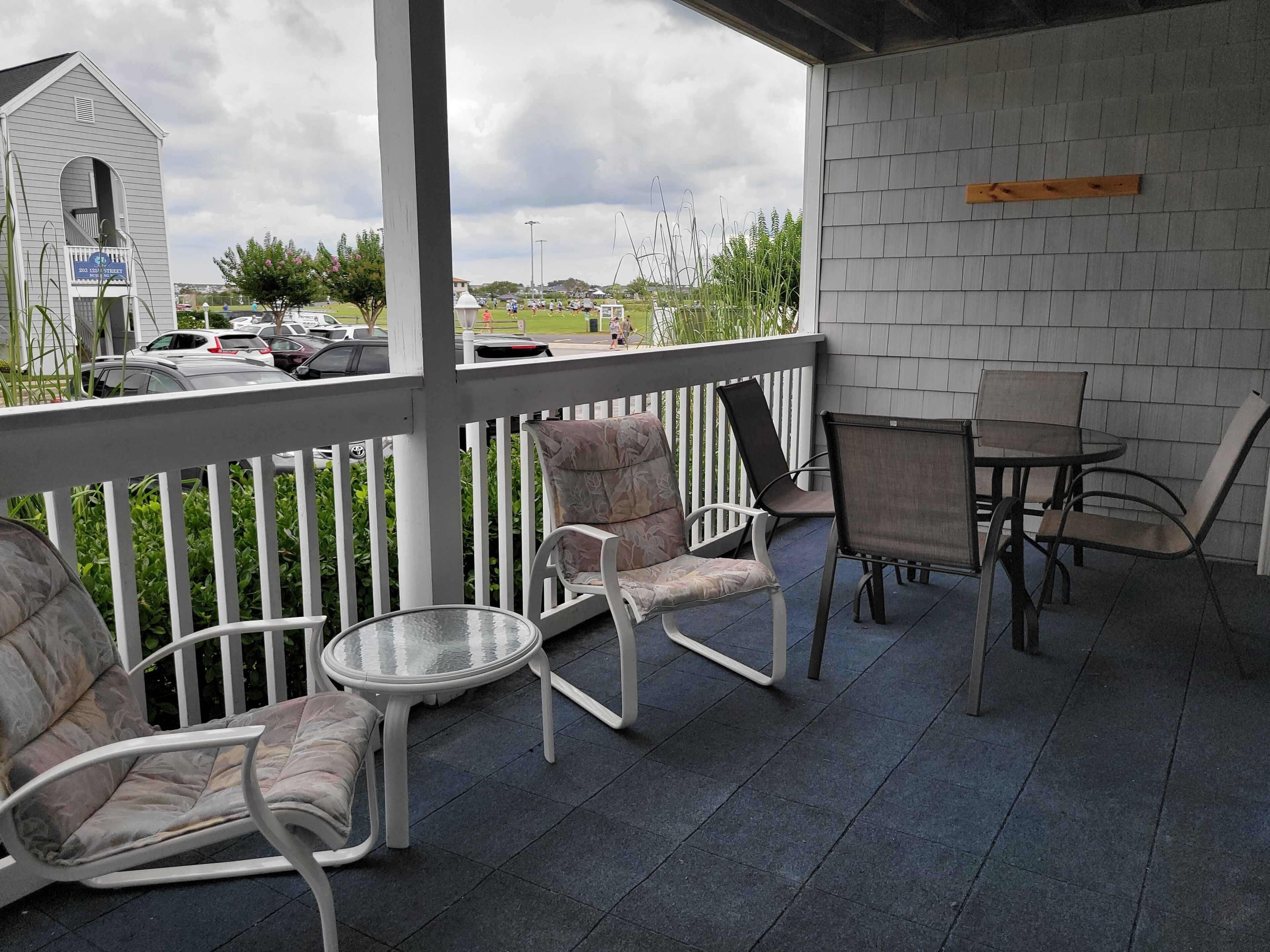 Un espace extérieur abrité, avec des chaises et une table, une clôture blanche, et une vue sur les voitures garées et la verdure en arrière-plan.